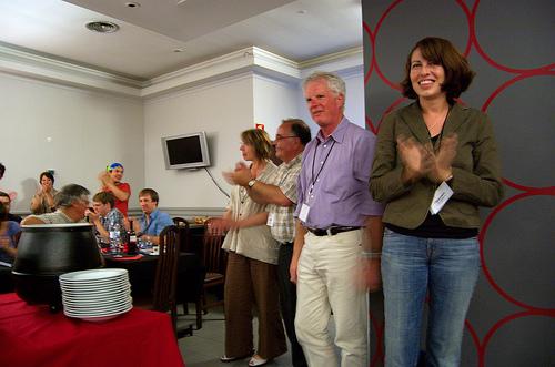 Four people standing in front, with people sitting in a restaurant setup; everybody is clapping their hands