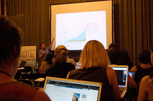 Attendees, some with laptops, listening to a presentation on stage, with a graph projected to the screen