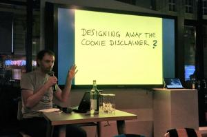 A person sitting behind a table on a stage, with a bright yellow slide displaying the title 'Designing away the cookie disclaimer', followed by a question mark
