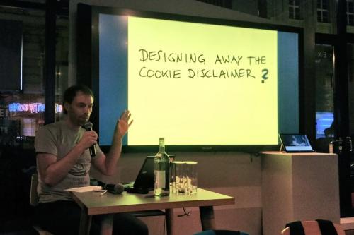 A person sitting behind a table on a stage, with a bright yellow slide displaying the title 'Designing away the cookie disclaimer', followed by a question mark