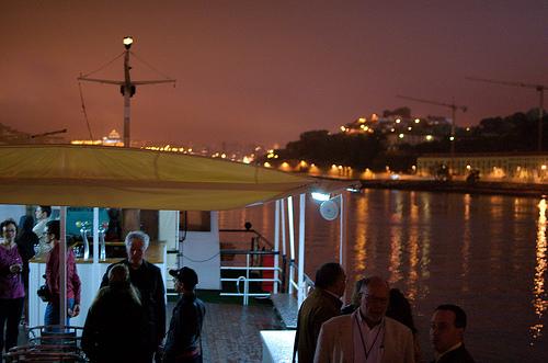 People standing on a river cruise boat chatting, with a cityscape and an illuminated sky behind