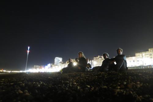Four people sitting on a pebbled beach, with a bright front of houses in the distance