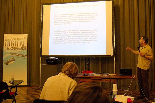 The speaker on stage, in front of a screen with a slide full of text