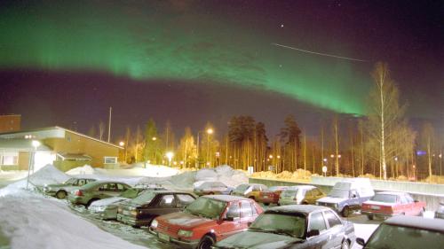 A strong display of green aurora borealis over a snowy parking lot with cars, illuminated by street lights.