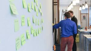 A whiteboard with post-it notes, two persons in the background engaged in a discussion